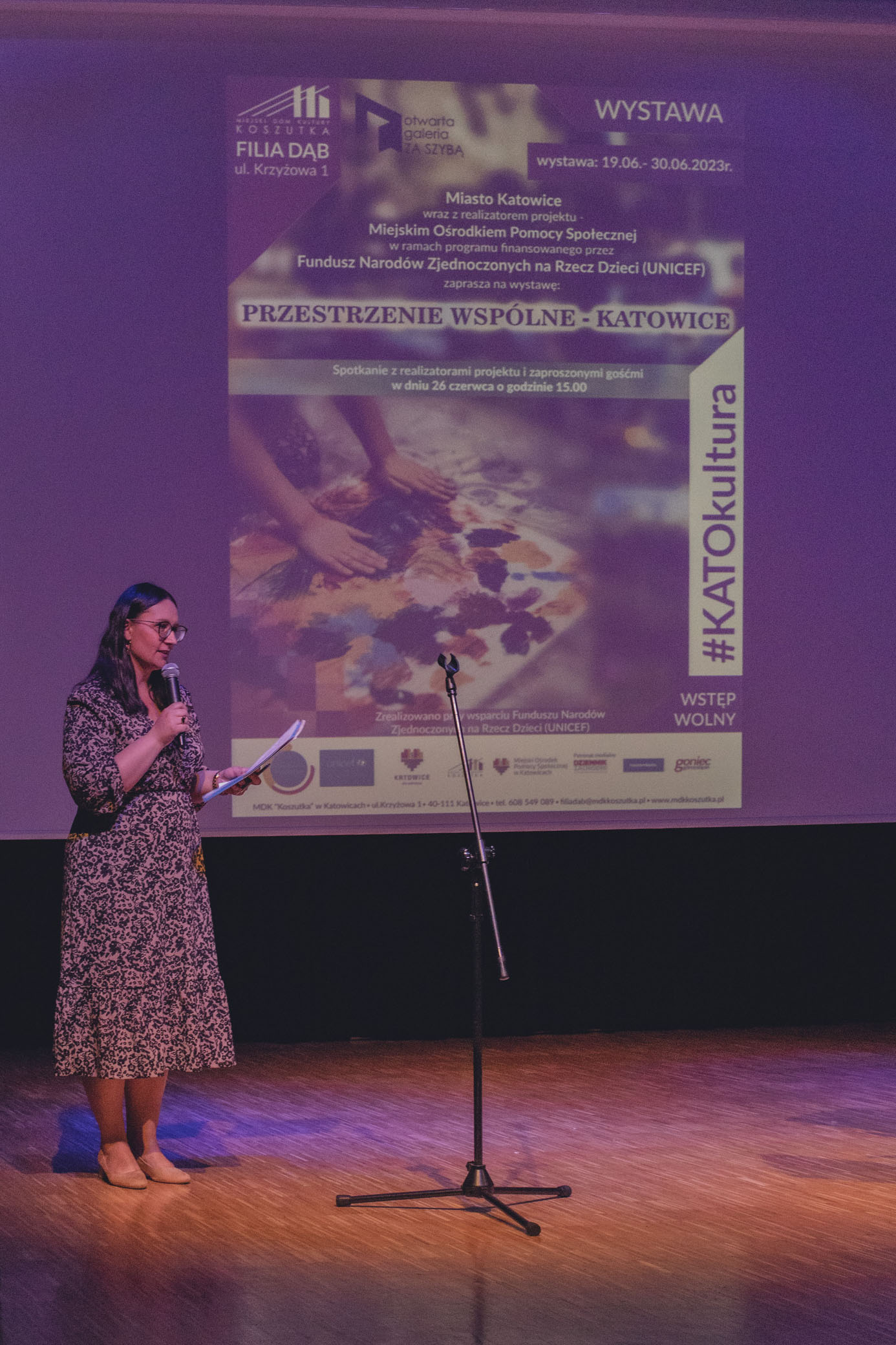 Zdjęcie przedstawia kobietę prowadzącą spotkanie. Kobieta stoi na scenie i mówi do mikrofonu. W tle znajduje się plakat informujące o wystwie "Przestrzenie wspólne - Katowice".