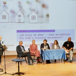 Na scenie widać siedzące przy stole osoby patrzące na mówiącego mężczyznę przy konferansjerce. W tle na ekranie widać nazwę konferencji oraz logo.