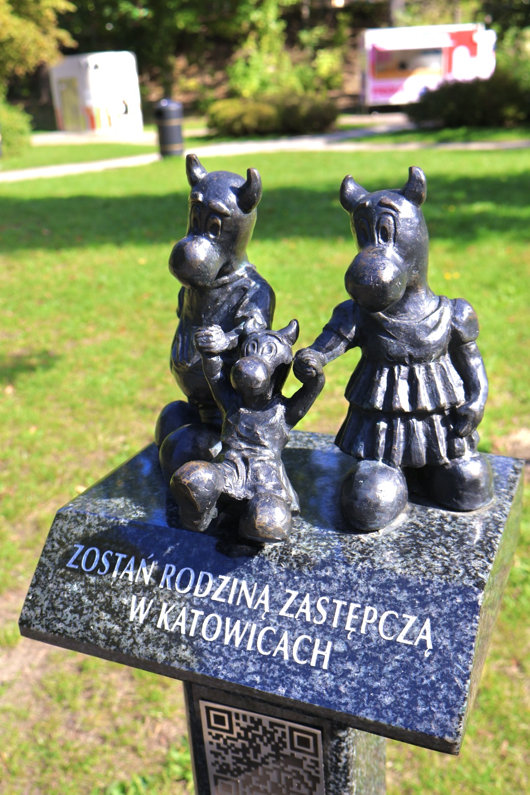 Rzeźba przedstawiająca rodzinę katowickich Beboków na tle parku z napisem na tabliczce „Zostań rodziną zastępczą w Katowicach”.